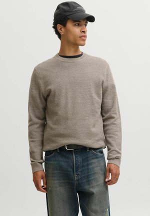 Jeune homme portant un pull en maille gris, un jean bleu foncé, une ceinture noire et une casquette noire, regardant vers la droite sur un fond clair uni.