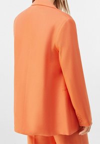 Oranžový blazer s klopovým límcem, strukturovanými rameny, jedním rozparkem vzadu a knoflíkovými manžetami. Hladká textura a střihové provedení.