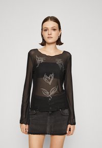 Svart genomskinlig långärmad topp med blommig broderi och metalliska detaljer, med en vågig halsringning och en knappdetalj vid fållen.