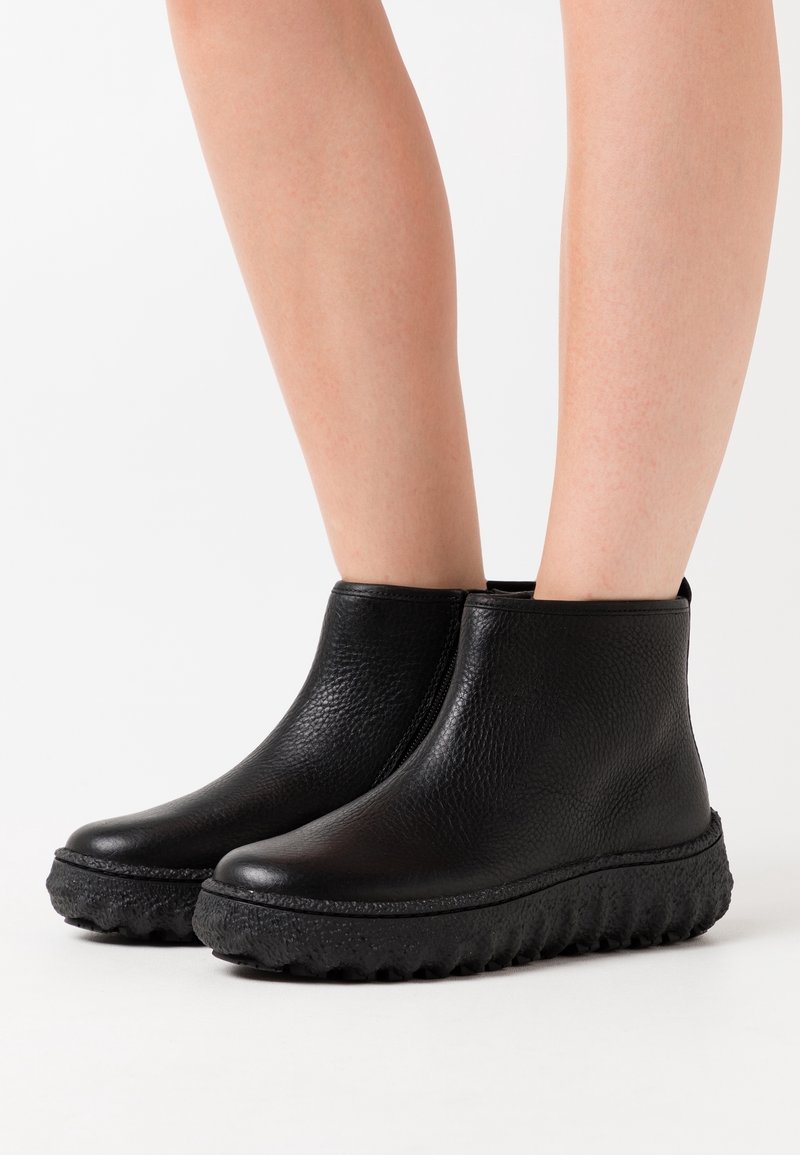 Jambes portant des bottines en cuir noir texturé avec des semelles épaisses et robustes sur un fond blanc uni.