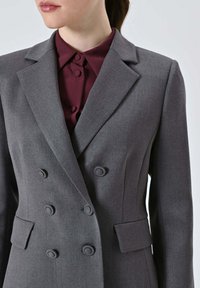 Blazer grigio doppiopetto con vestibilità sartoriale. Presenta rever con intagli, due tasche frontali e bottoni rotondi neri; tessuto dalla consistenza liscia.