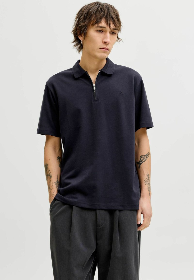 Junger Mann mit mittellangem Haar, der ein schwarzes Poloshirt mit kurzem Reißverschluss und dunkelgraue Hose trägt und vor einem einfarbigen Hintergrund steht.