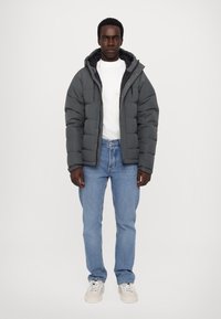Doudoune grise avec capuche, fermeture éclair devant et doublure noire, associée à un jean bleu clair et des baskets blanches. Design simple, détails fonctionnels.