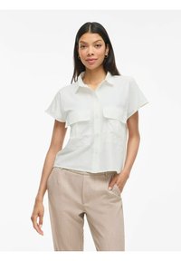 Lichtgekleurde shirt met korte mouwen en kraag, voorzien van twee borstzakken, knoopsluiting en een relaxed fit. Gepaard met beige broek.