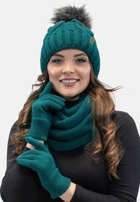 Bonnet tricoté bleu sarcelle avec pompon, écharpe assortie et gants. L'écharpe présente une texture tissée ; les gants ont des poignets côtelés.