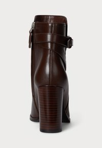 Bota de tornozelo em pele castanha com fecho de zip e um detalhe de tira curvada. O salto é quadrado e com textura de grão de madeira.
