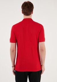 Červené polo tričko se strukturovaným límcem, krátkými rukávy a hladkým povrchem. Jednoduchý design bez viditelných vzorů nebo ozdob.