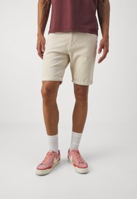 Beige farkkushortsit, joiden lahkeet on käännetty, yhdistetty vaaleanpunaisiin lenkkareihin ja valkoisiin nilkkasukkasiin, päällä maroon-punainen t-paita.