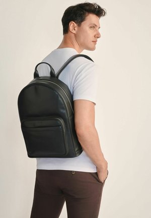 Czarny skórzany plecak o zaokrąglonym kształcie, z przednią kieszenią i podwójnymi paskami na ramiona. Gładka faktura, z subtelnym detalem logo.