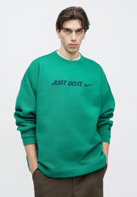 Πράσινο φούτερ Nike με στρογγυλή λαιμόκοψη, κεντημένο κείμενο "JUST DO IT." σε ναυτικό μπλε και λογότυπο της Nike στο μανίκι. Πυκνό, υφασμένο ύφασμα.
