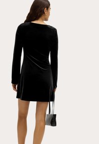 Schwarzes Samtkleid mit langen Ärmeln und taillierter Silhouette sowie dezentem Fall, kombiniert mit einer kleinen schwarzen Handtasche mit elegantem Design.