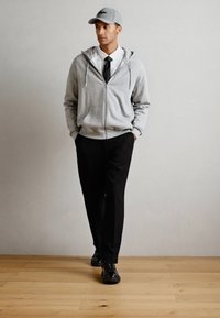 Camisola cinza com fecho, sobre uma camisa branca e gravata preta, combinada com calças pretas e ténis pretos, finalizada com um boné cinza.