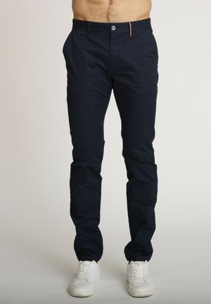 Homme portant un pantalon slim bleu marine foncé et des baskets blanches, debout devant un fond clair uni.