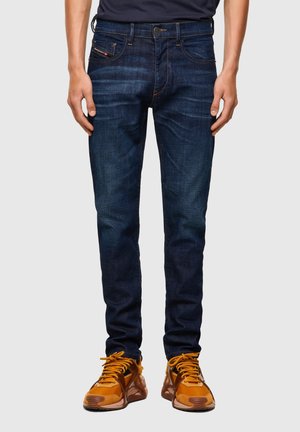 Schmal geschnittene dunkelblaue Jeans, kombiniert mit orange-braunen Sneakern, dargestellt von der Taille bis zu den Füßen vor einem einfarbigen Hintergrund.