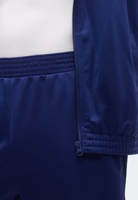 Nærbilde av marineblå glidelåsjakke og bukser med elastisk midjebånd på en person iført en hvit topp.