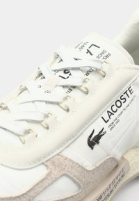 Witte sneaker met grijze accenten, textuurmaterialen, opvallende stiksels en gedrukt merklogo. Beschikt over een traditioneel vetersysteem en logo.