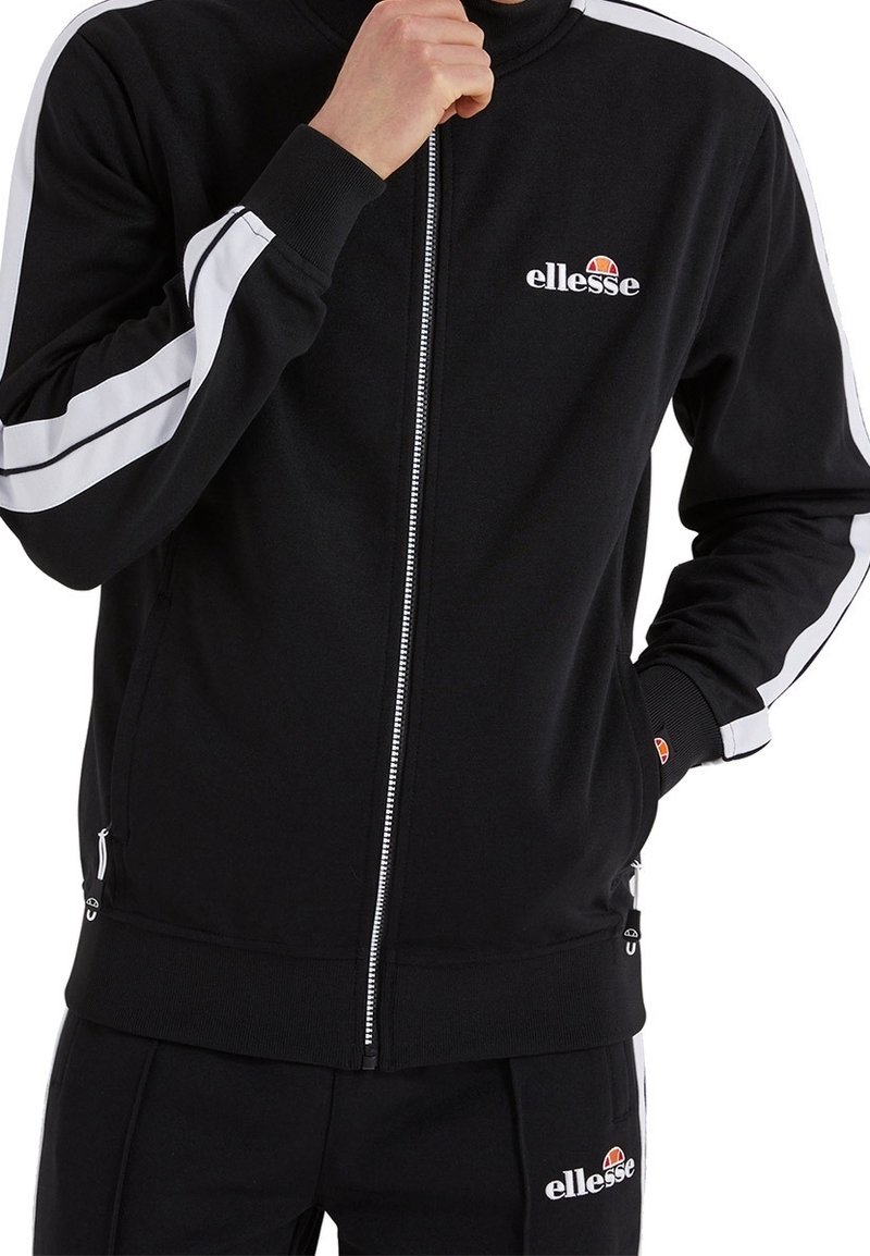 Svart zip-up träningsoveralljacka med vita ränder på axlarna, ribbade ärmslut och krage. Har en logotyp på vänster bröst och dragkedjefickor.