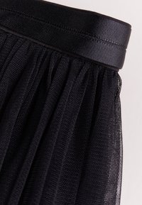 Gros plan sur un tissu en maille noire rassemblé, attaché à une ceinture élastique noire large et texturée, sur un fond neutre.