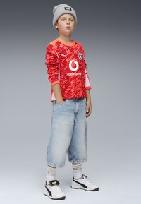 Pitkähihainen punaisia kuvioita sisältävä paita, jossa on valkoisia yksityiskohtia, yhdistettynä vaaleansinisihin löysiin farkkushortseihin ja valkoisiin lenkkareihin, asusteena harmaa pipo.