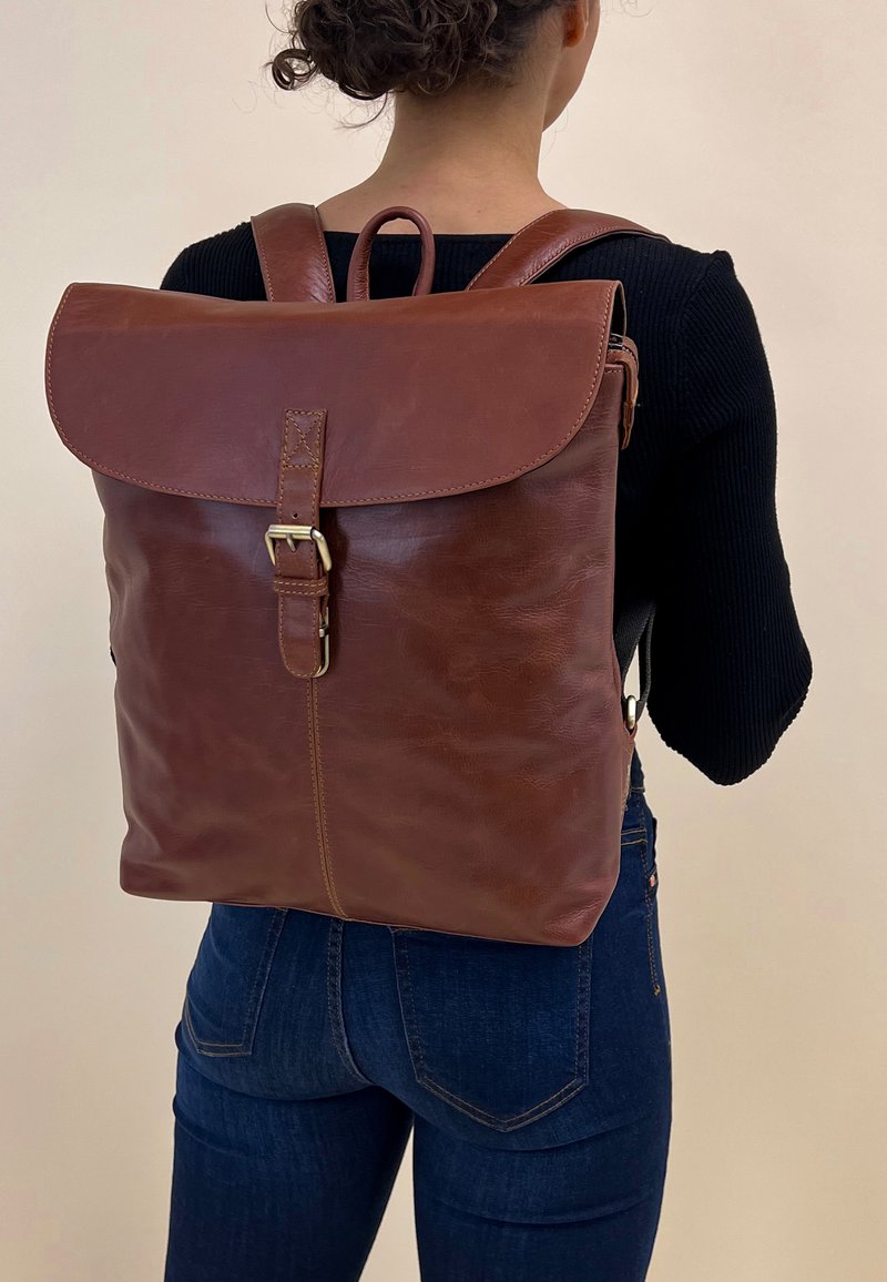 Sac à dos en cuir marron avec une fermeture à rabat, des bretelles réglables et une boucle à l'avant. Texture lisse et design minimaliste.