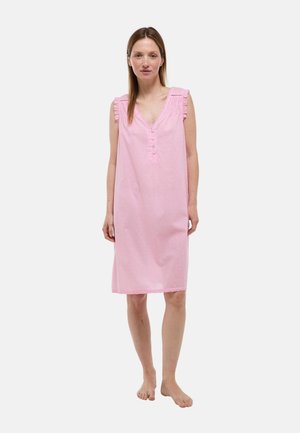 Femme debout pieds nus portant une robe rose sans manches aux genoux avec des boutons et des détails à volants aux épaules sur un fond uni.