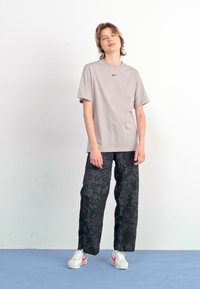 Ανοιχτό γκρι T-shirt με λογότυπο, χαλαρό μαύρο παντελόνι με λουλούδια και λευκά αθλητικά παπούτσια με κόκκινες λεπτομέρειες, στέκεται μπροστά από ένα απλό φόντο.
