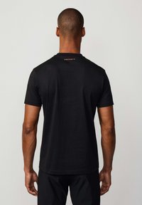 Camiseta de algodón negra con cuello redondo y mangas cortas. Un logotipo bordado en naranja está situado en la parte superior de la espalda.