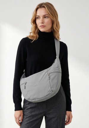 Donna con dolcevita nero che indossa una grande borsa a tracolla grigia con più cerniere e logo "GMV", in piedi davanti a uno sfondo semplice.