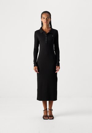 Abito nero a maniche lunghe con colletto polo, bottoni e silhouette aderente. Tessuto morbido, lunghezza fino al polpaccio, abbinato a sandali con cinturini.