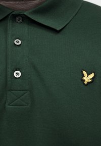 Πράσινο σκούρο πόλο μπλουζάκι με μαύρα κουμπιά και μικρό κίτρινο έμβλημα αετού κεντημένο στην αριστερή πλευρά στο στήθος.