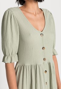 Jasnozielona sukienka o teksturze z bufkami, falbaniastymi mankietami, dekoltem w serek oraz guzikami w kolorze żółwia z przodu, noszona przez kobietę.
