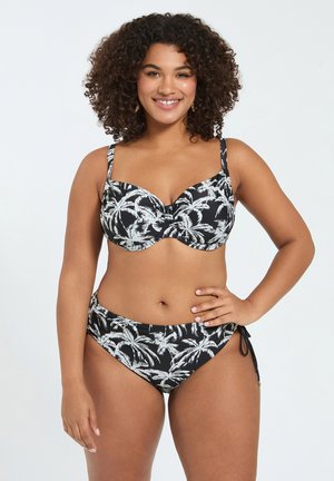 Kiharatukkainen nainen hymyilee, pukeutuneena mustavalkoiseen palmukuosiseen bikiniin, jossa on säädettävät sivusolmukkeet, seisoo yksiväristä taustaa vasten.