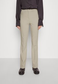 Persoon in beige rechte broek, zwart langemouwen shirt en zwarte enkelboots, staand tegen een effen lichte achtergrond.