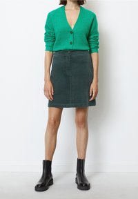 Grüne Strickjacke mit V-Ausschnitt und Knopfverschluss, kombiniert mit einem dunkelgrünen Rock. Schwarze Stiefeletten runden das Outfit ab.