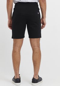 Fekete pamut shorts, elasztikus derékbanddal és hátsó zsebből, testhez álló kialakítással és sima textúrával, fekete sportcipővel viselve.