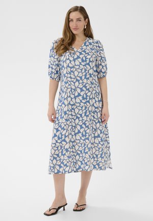 Kobieta w niebieskiej sukience midi z białym wzorem liści, z bufkami i czarnych sandałach na obcasie, stojąca na jednolitym tle.