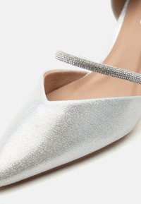 Ασημί μπαλαρίνα με μυτερή μύτη, λαμπερή υφή και λεπτό λουράκι διακοσμημένο με μικρές στρας στο επάνω μέρος.
