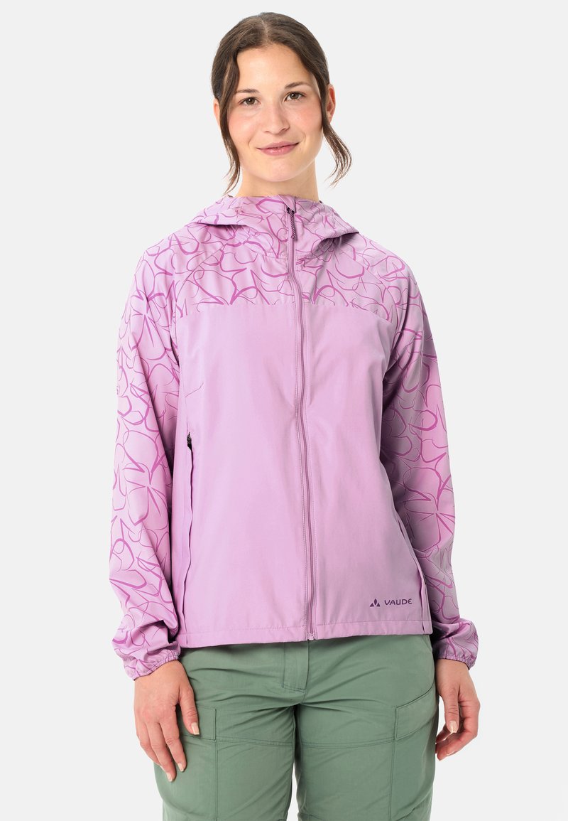 Femme portant une veste à capuche violette claire avec des motifs abstraits sur les manches et le haut de la poitrine, associée à un pantalon cargo vert.