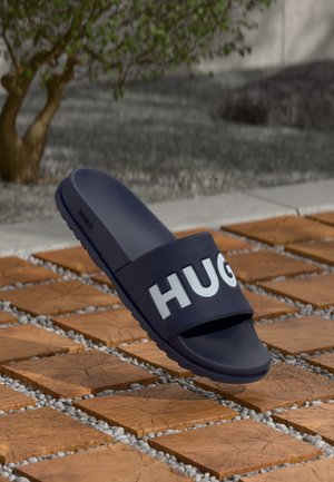 Tengerészkék, gumiból készült papucs széles pánttal, amelyen a "HUGO" felirat fehér betűkkel szerepel, texturált talppal és lapos talppal.