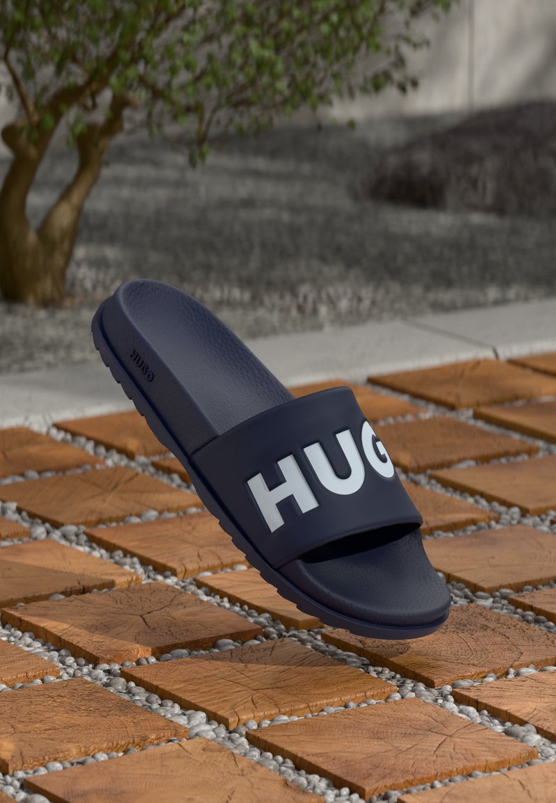Jūrų laivyno guminių šlepečių su plačia juosta, ant kurios balta spalva parašyta "HUGO", dizainas su tekstūruotu pado paviršiumi ir lygia sola.