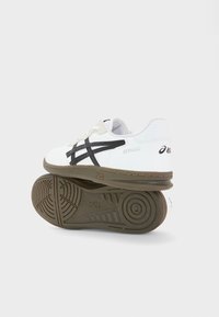 Chaussures de sport blanches avec une tige en cuir texturé, des rayures noires et une semelle en caoutchouc marron à motif. Le talon est marqué par un logo.