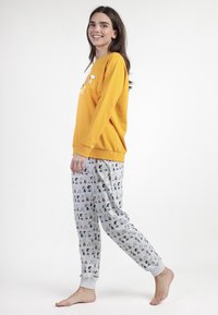 Joven sonriendo, vistiendo una camiseta de manga larga naranja y pantalones de pijama grises con estampados de personajes de dibujos animados, caminando descalza sobre un fondo blanco.