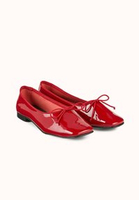 Ballerines en cuir verni rouge avec un design à bout pointu, présentant un détail de nœud à l'avant et un petit talon noir. Texture lisse avec une finition brillante.