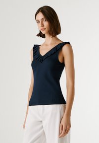 Marinblå ärmlös topp med ribbad textur, har en V-ringning prydd med volangkant längs axlarna. Kombinerad med ljusa byxor.