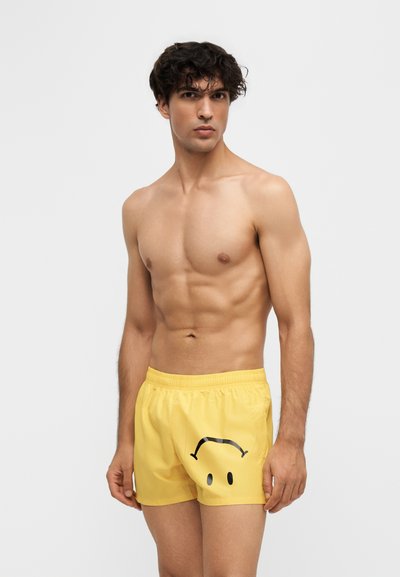 Jeune homme torse nu aux cheveux bouclés foncés portant un short jaune avec un smiley noir froncé, debout devant un fond uni.