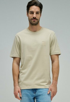 Hombre con cabello oscuro y barba, vestido con una camiseta beige de manga corta y jeans azul claro, frente a un fondo gris liso.