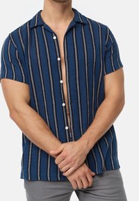 Niebieska koszula z krótkim rękawem, ozdobiona pionowymi czarno-białymi paskami, z zapięciem na guziki i luźnym krojem, wykonana z teksturowanego materiału.