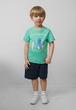Zielony bawełniany t-shirt z grafiką deskorolek w różnych kolorach i stylach; zestawiony z ciemnymi szortami i białymi trampkami.