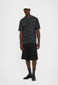 Man standing wearing black EA7 logo print t-shirt, black wide shorts, dark socks, black shoes, and a white cap on a plain white background.
