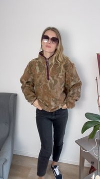 Pullover de fleece castanho e bege cortado, com padrão abstrato e fecho roxo, combinado com calças pretas e sapatilhas pretas.